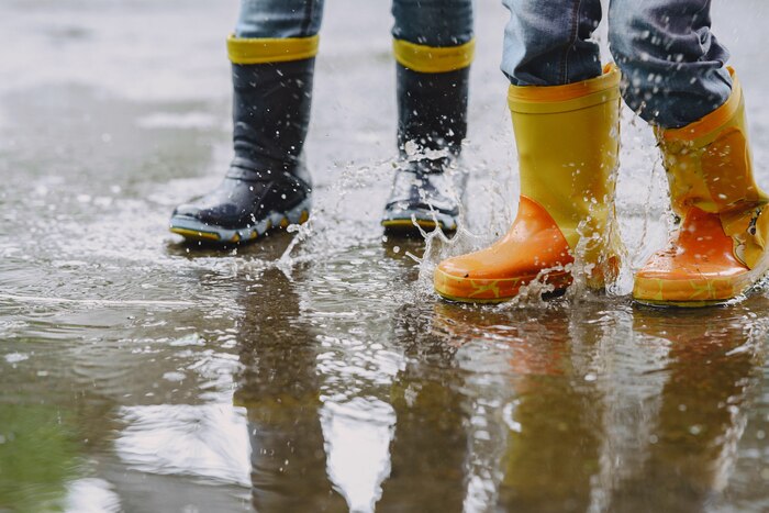 Funny kids in rain boots playing with paper ship by a puddle