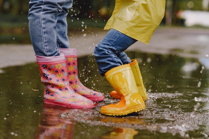 Funny kids in rain boots playing with paper ship by a puddle