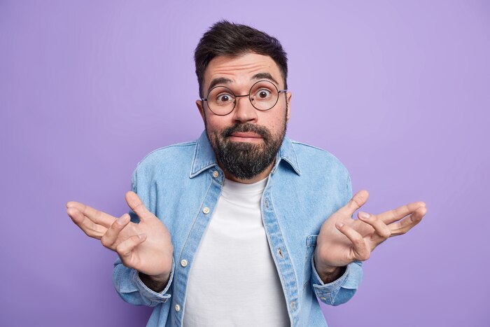 Man spreads palms feels clueless and uncertain canot make choice wears round eyeglasses denim shirt poses